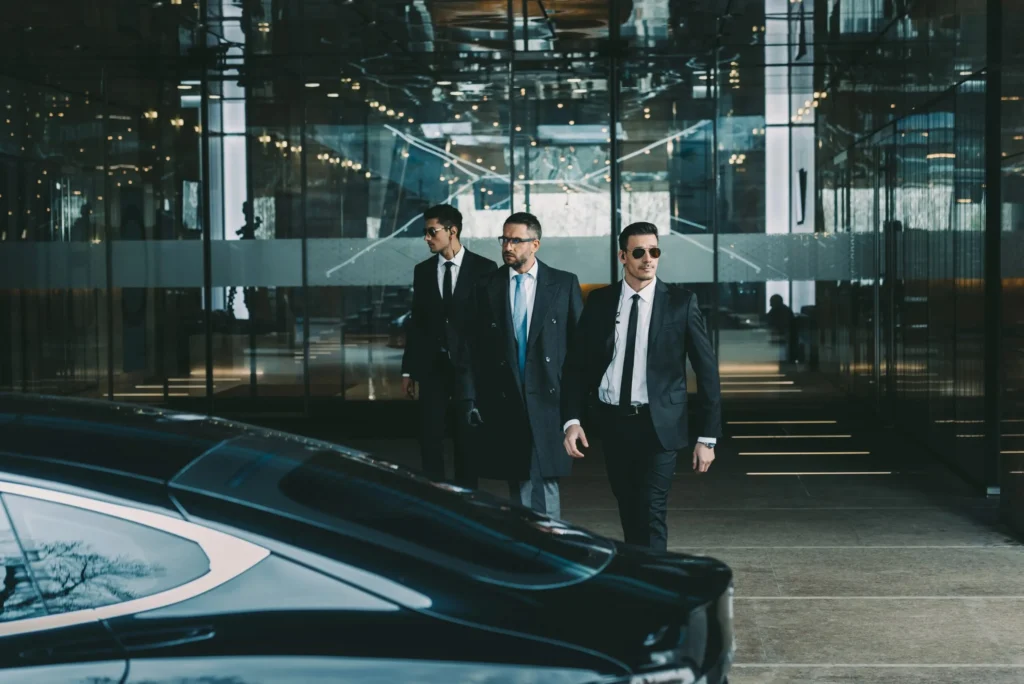 Close protection security team escorting an executive from a modern commercial building towards a waiting vehicle.