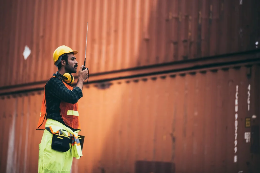 Industrial worker in high-visibility PPE using a handheld two-way radio beside shipping containers.