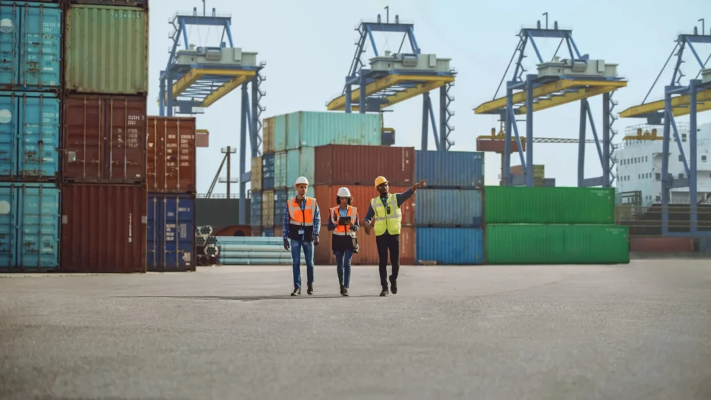 Port workers in high-visibility PPE coordinating operations among shipping containers and cargo cranes.